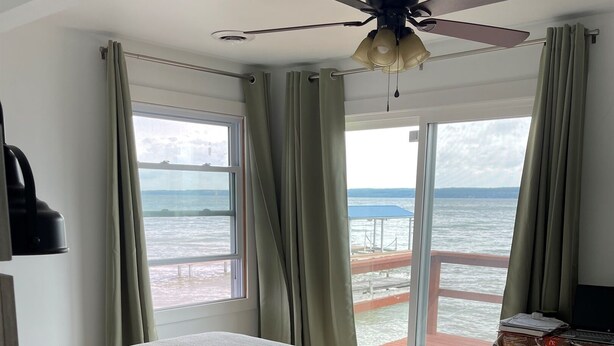 Un dormitori amb un llit, un ventilador de sostre, una finestra amb cortines i vista al mar.