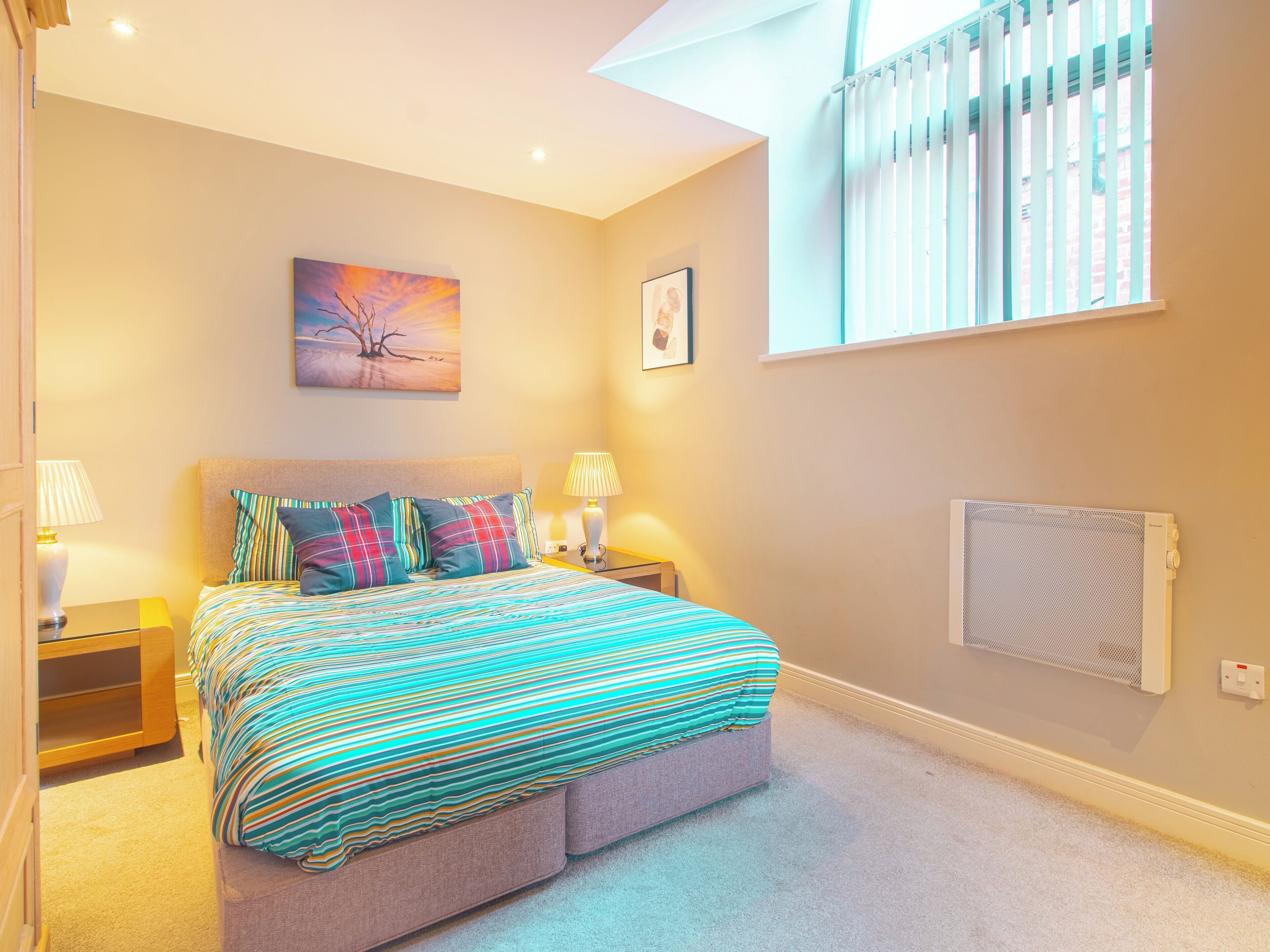 Une chambre à coucher comprenant un lit, deux tables de chevet, une fenêtre avec des stores, un radiateur et un tableau accroché au mur.