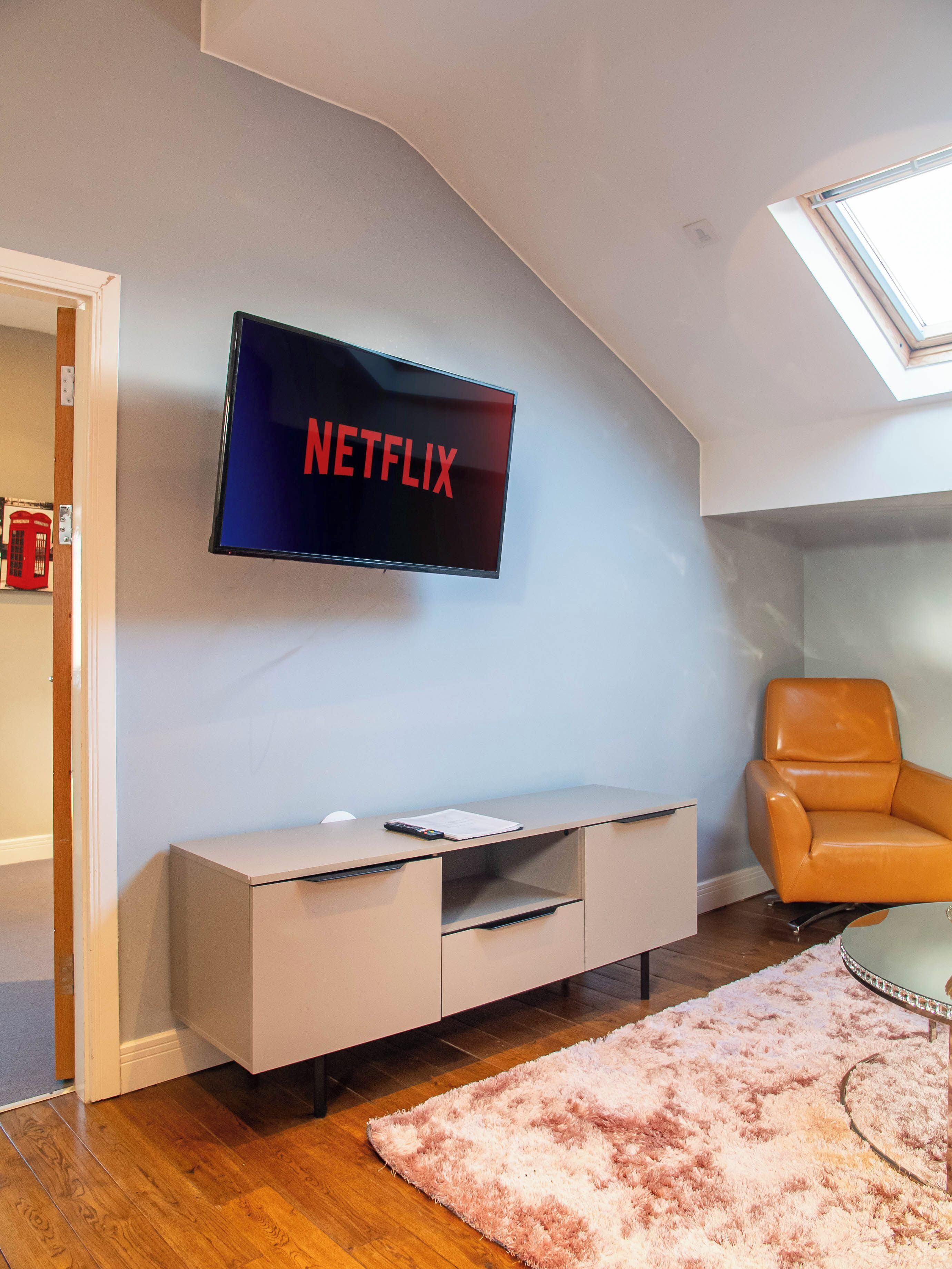 A modern living room with a mounted TV displaying "NETFLIX," a beige entertainment unit, a brown leather chair, and a skylight.