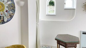 A room with a circular mosaic wall art, a wicker chair, two arched windows, and a white wall. - Villa Sain Charming Suite (Anacapri)