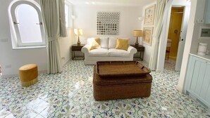 A living room with a white sofa, two side tables with lamps, a patterned floor, and a large window with curtains. - Villa Sain Charming Suite (Anacapri)