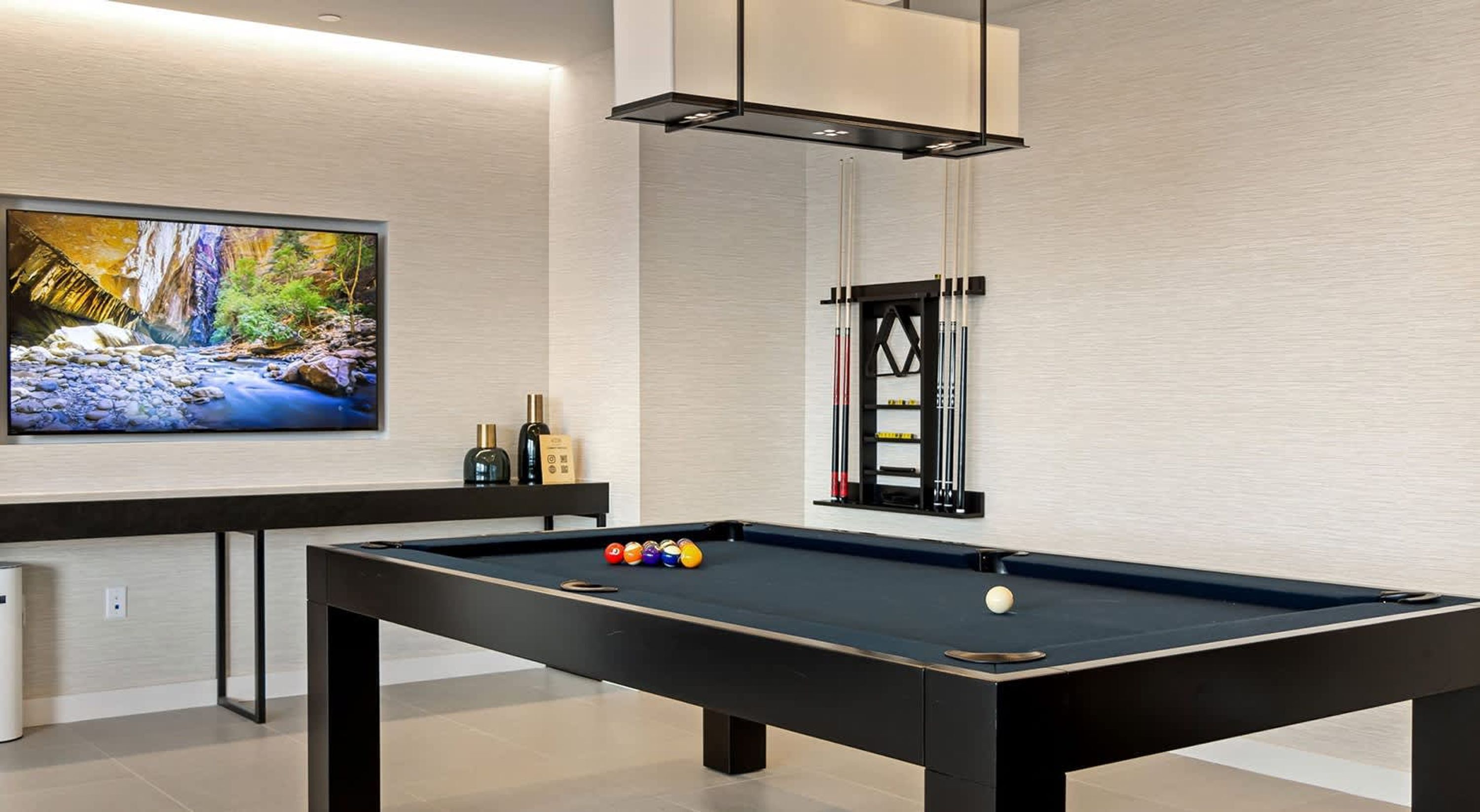 Une table de billard moderne, avec un porte-billes et des queues de billard, un téléviseur diffusant des images de la nature, et une petite étagère avec des bouteilles.