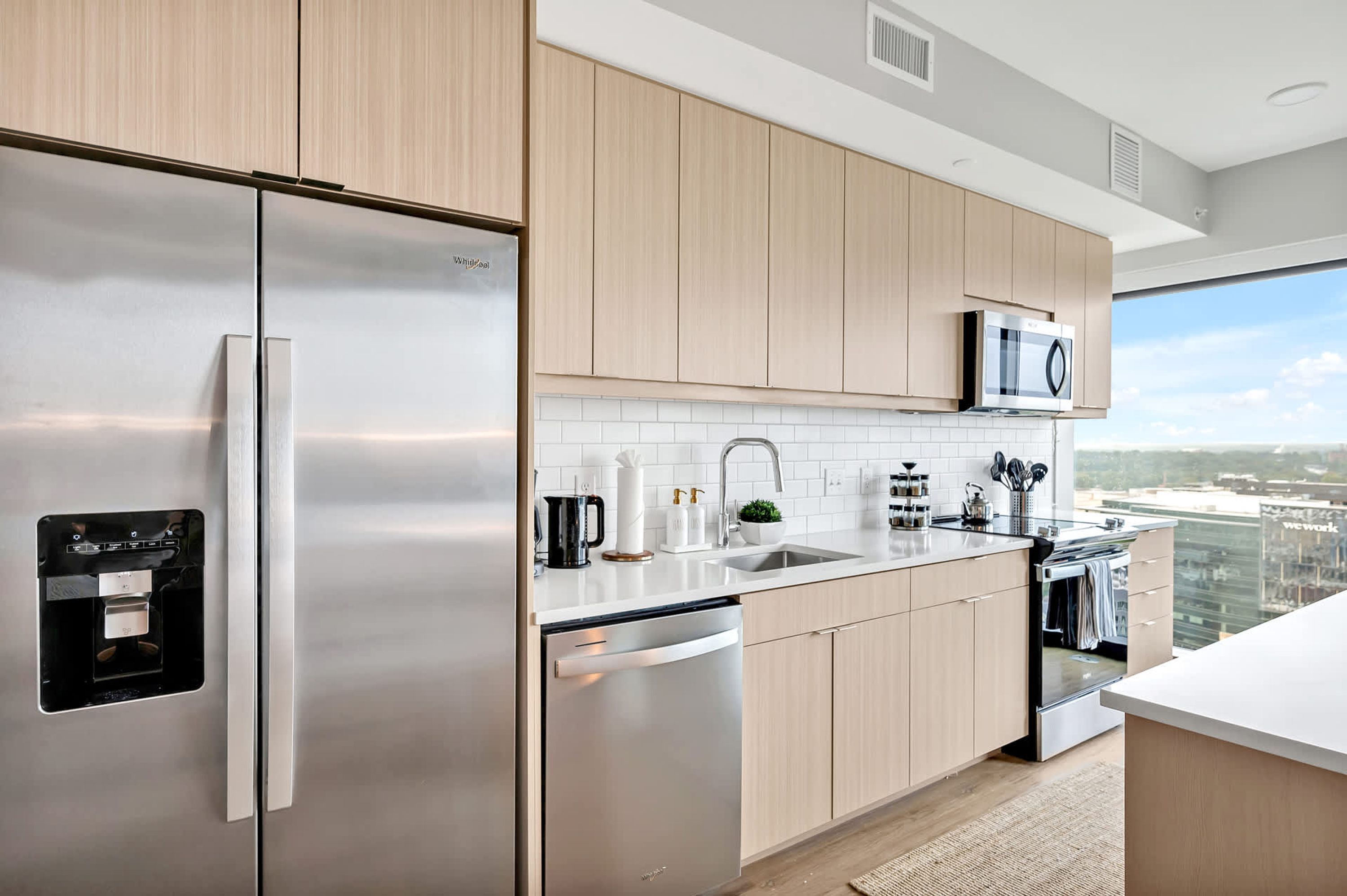 A modern kitchen with stainless steel appliances, wooden cabinets, and a large window offering a city view.