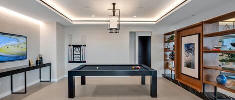 Une table de billard moderne dans une pièce bien éclairée, avec un téléviseur à écran plat, des étagères remplies de livres et d’objets décoratifs, et un luminaire au plafond.