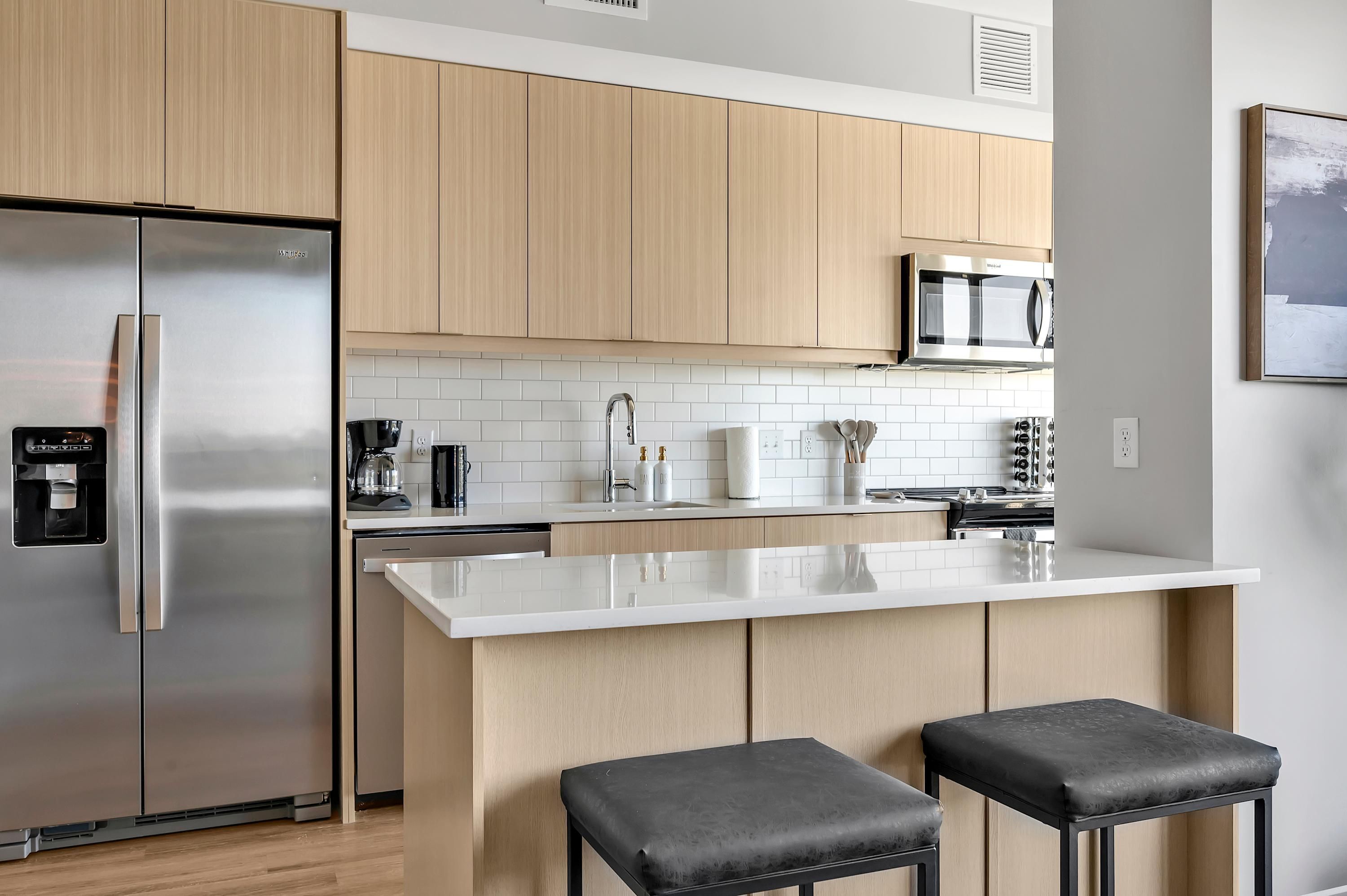 Een moderne keuken met lichte houten kastjes, roestvrijstalen apparatuur, een wit aanrecht en twee zwarte barkrukken.