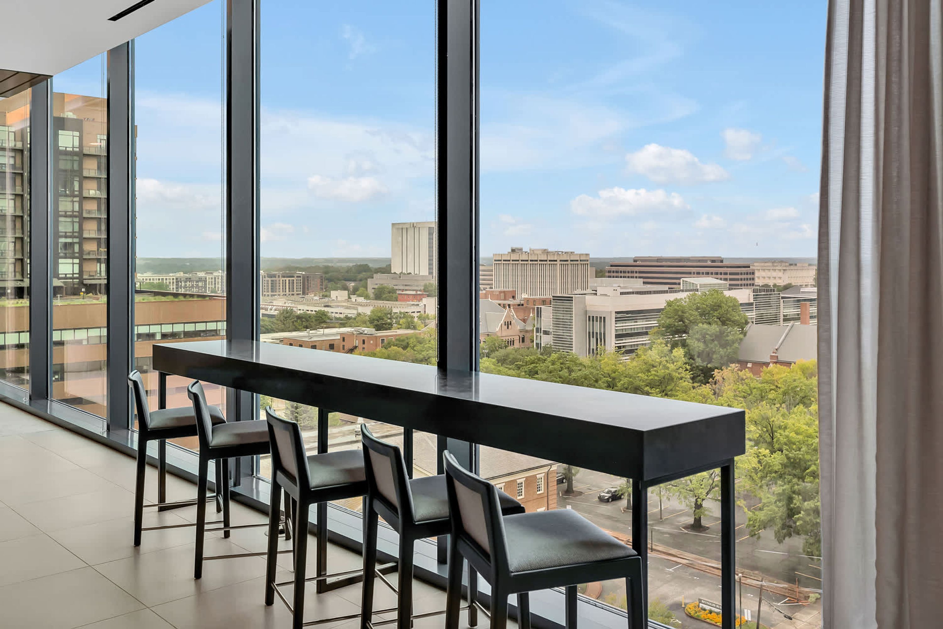 A modern room with a long table and four chairs, large windows offering a city view.