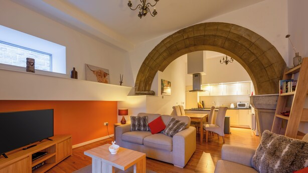 Sala de estar con televisor, sofá y mesa de centro. Se observa una cocina al fondo con zona de comedor.