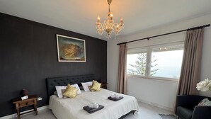 A modern bedroom with a large bed, a chandelier, a painting, and a view of the outdoors.