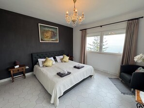 A modern bedroom with a large bed, a chandelier, a painting, and a view of the outdoors.