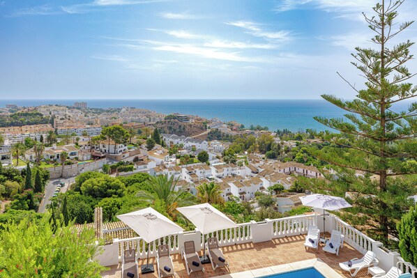 A coastal residential area with a view of the sea, white buildings, and a pool area with sun loungers.