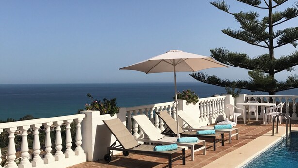 A poolside area with lounge chairs and an umbrella, overlooking a clear blue sky and the ocean.