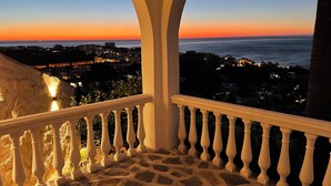 A balcony with a stone floor and white balustrade, overlooking a coastal cityscape during sunset.