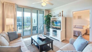 Amplia sala de estar con sofá, mesa de centro y mueble para la televisión. Cuenta con un balcón con vistas a edificios y un ventilador de techo.