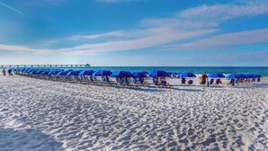 Una playa con hileras de sombrillas azules y sillas, un muelle al fondo y cielo despejado.