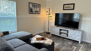 Ein Wohnzimmer mit einer grauen Eckcouch, einem Flachbildfernseher auf einem weißen Schrank, einer Stehlampe und zwei gerahmten Bildern an der Wand.