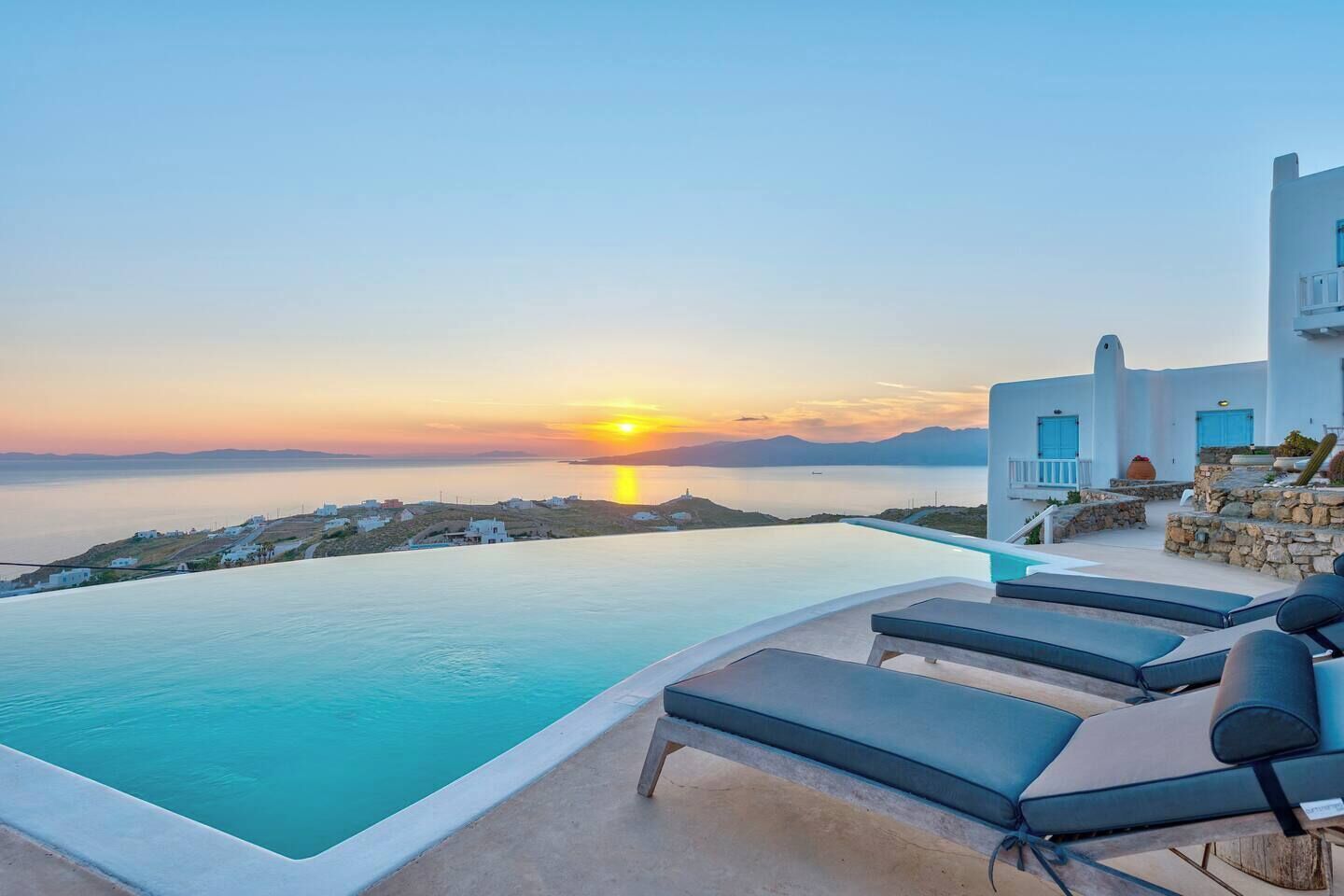 An infinity pool with a view of the sunset over the sea, a white building with blue doors, and a stone wall with plants.