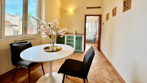 A dining area with a round table, two chairs, a vase with flowers, a kitchen in the background, and a view of the outside. - Terrace Studio   Apartment with panoramic terrace (Florence)