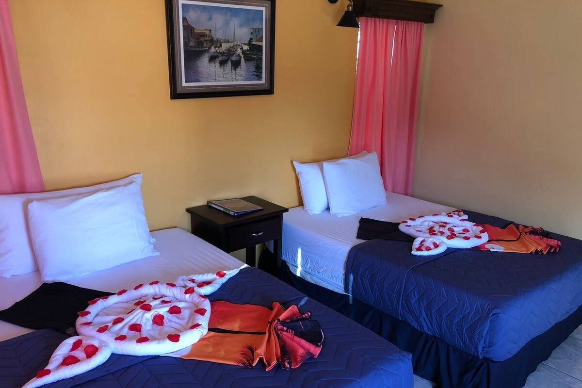 Habitación de hotel con dos camas, cada una con cubrecama azul y almohadas blancas. Se observa una mesita de noche con un libro y una lámpara, además de un cuadro en la pared.