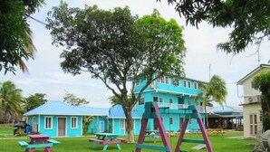 Un parque infantil con columpios y mesas de picnic en un espacio verde, con un edificio azul al fondo.