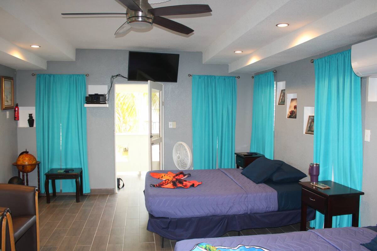 Habitación de hotel con dos camas, ventilador de techo, televisor y ventana con cortinas.