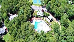 Vista aérea de una propiedad con cancha de tenis, piscina y varios edificios rodeados de árboles densos.