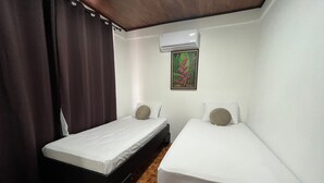 Een kleine, schone kamer met twee eenpersoonsbedden, een houten vloer, een plafondventilator en een wandairconditioner.