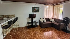 Een woonkamer met een bank, een televisie, een ventilator en een eethoek met stoelen en een tafel.