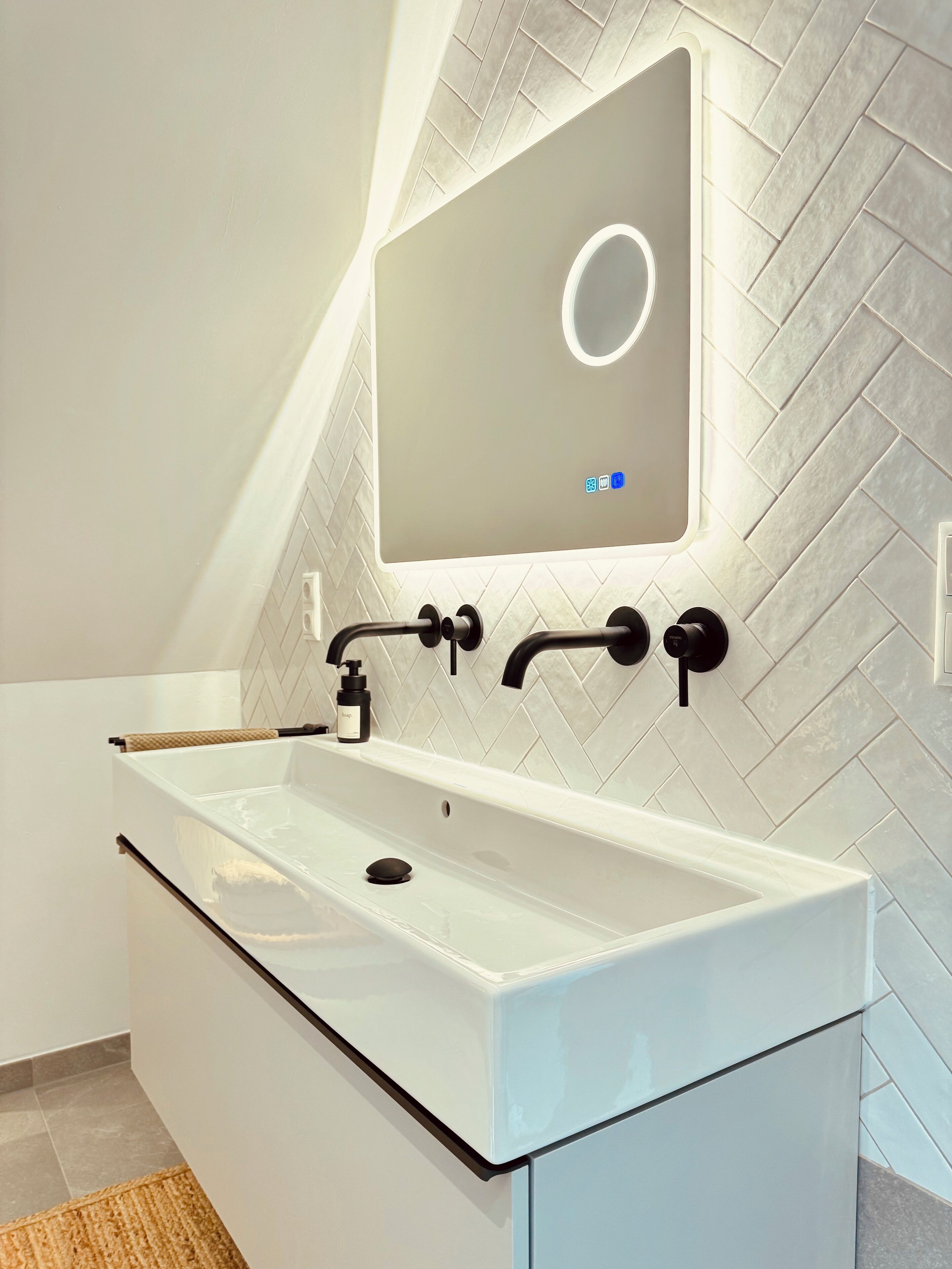 A modern bathroom with a white vanity, a large mirror, and a herringbone-patterned wall.