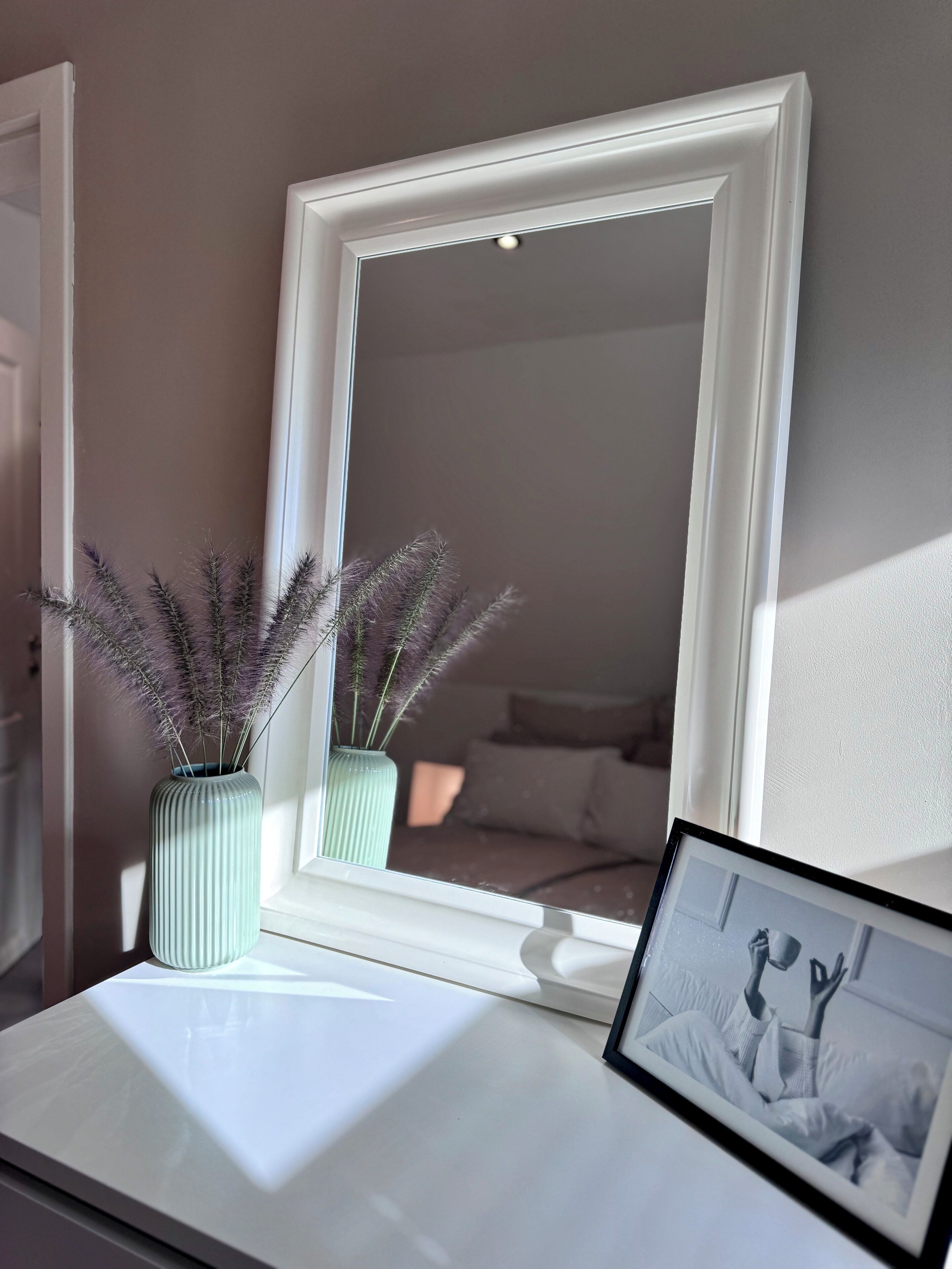 A mirror with a white frame on a white surface, reflecting a room with a bed and pillows. There are two vases with dried plants on the surface.