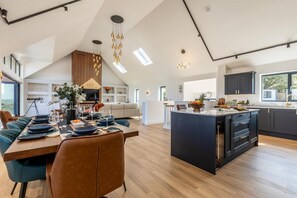 Une cuisine moderne avec un îlot central, un coin repas doté d’une longue table et une chambre en mezzanine avec une grande fenêtre.