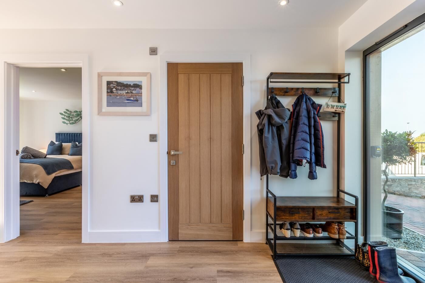 Um interior moderno com uma porta de madeira, um cabideiro com jaquetas, uma cama com cabeceira azul e um quadro emoldurado na parede.