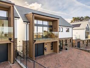 Une maison moderne à deux étages, dotée d’une terrasse en briques, de grandes fenêtres et d’un balcon avec des garde-corps en verre.