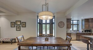 Un espace repas comprenant une table en bois, des chaises, un lustre et une vue dégagée sur l’extérieur grâce à de grandes fenêtres.