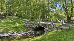 Un pont de pierre enjambe un ruisseau dans une zone boisée, avec une route pavée en arrière-plan.
