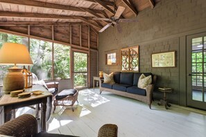 Une pièce spacieuse avec un plafond en bois, un canapé avec des coussins, une table avec une lampe et une fenêtre donnant sur des arbres.