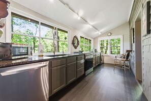 Een keuken met een roestvrijstalen koelkast, een magnetron, een spoelbak en een fornuis. Grote ramen zorgen voor daglicht en uitzicht op bomen.
