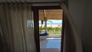 En åben dør, der fører ud til en balkon med udsigt over stranden, et palmetræ og udendørs møbler.