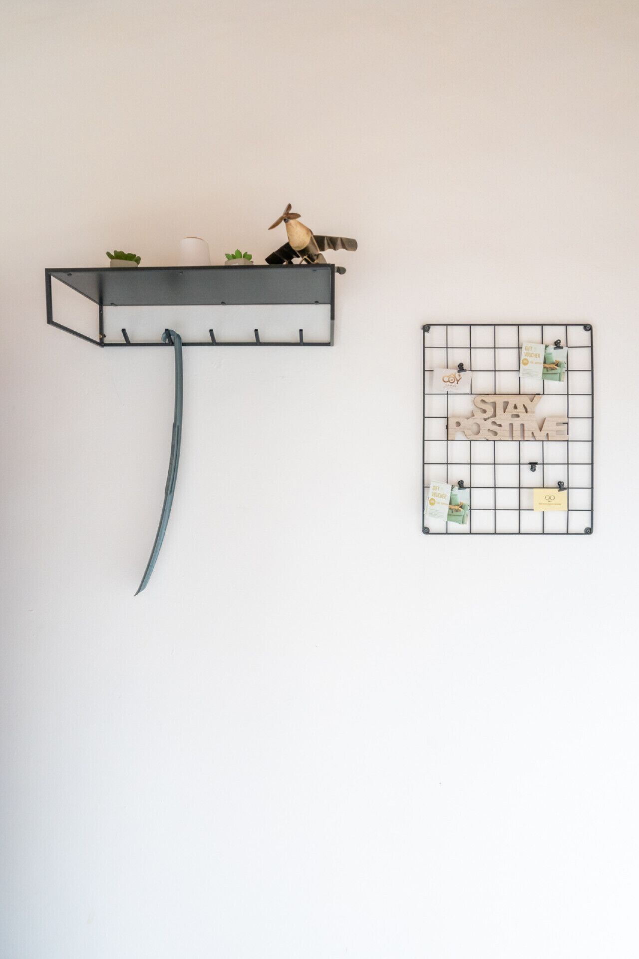 Eine Wandhalterung aus Metall mit einer nach unten gebogenen Metallstange, einer kleinen Vogelstatue und einem Gitterwandorganisator mit Karten und einem Holzschild.
