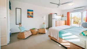 A bedroom with a bed, a ceiling fan, a window with curtains, a kitchen area, and wicker furniture. - Luxury studio, oasis of peace, centrally located close to most beautiful beaches (Sint Willibrordus)