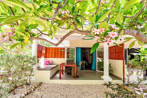 A covered outdoor seating area with a table and chairs, surrounded by lush greenery and flowering plants.
