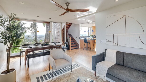 A modern living room with a dining area, a large window, a ceiling fan, and a staircase leading to an upper level.