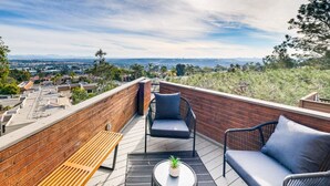 A rooftop terrace with a view of a cityscape, featuring a brick wall, a wooden bench, two wicker chairs, and a small table with a potted plant.