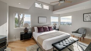 A modern bedroom with a large bed, a ceiling fan, and multiple windows offering a view.