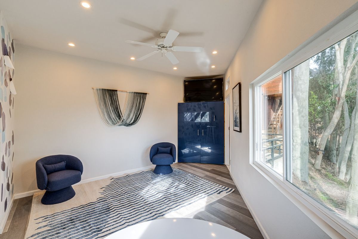 A modern room with a ceiling fan, a large window with a view of trees, a blue chair, and a black cabinet.