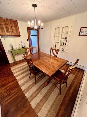 Une salle à manger avec une table et des chaises en bois, un lustre et un buffet vert où sont exposés des objets en verre.