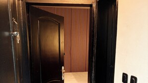 Un couloir étroit, avec un lit au fond, une porte à gauche et un mur blanc orné d’un panneau vertical.