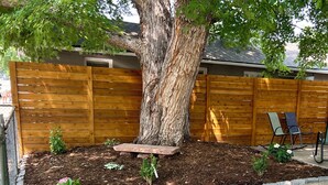 Un patio trasero con un árbol grande, una valla de madera, un banco de piedra y una zona con césped.
