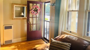 Una habitación con piso de madera, una puerta marrón con un adorno floral, una ventana con cortinas azules y un sofá de cuero marrón con almohadones a rayas.