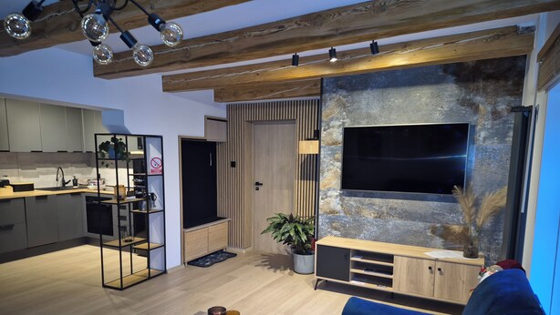 A modern living room with a large TV, wooden ceiling beams, a blue sofa, and a kitchen area with grey cabinets. - Apartamenty Rynek Szamotuły (Szamotuly)