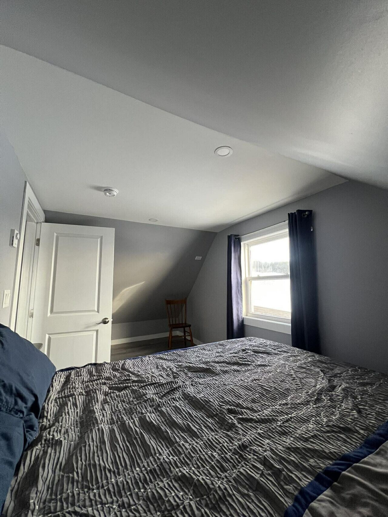 Un dormitorio con techo inclinado, una cama con colcha estampada, una silla, una ventana con cortinas y una puerta.